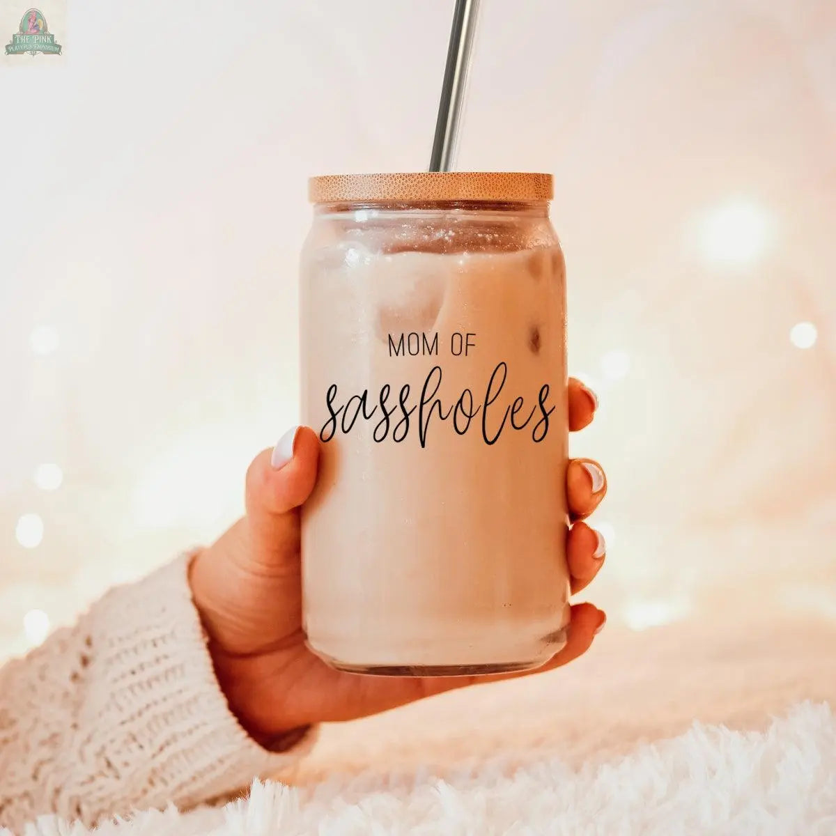 A hand holds the Mom of Sassholes 17oz glass cup with iced coffee, a bamboo lid, and a stainless steel straw. The person wears a cozy sweater, with softly glowing warm lights in the background.