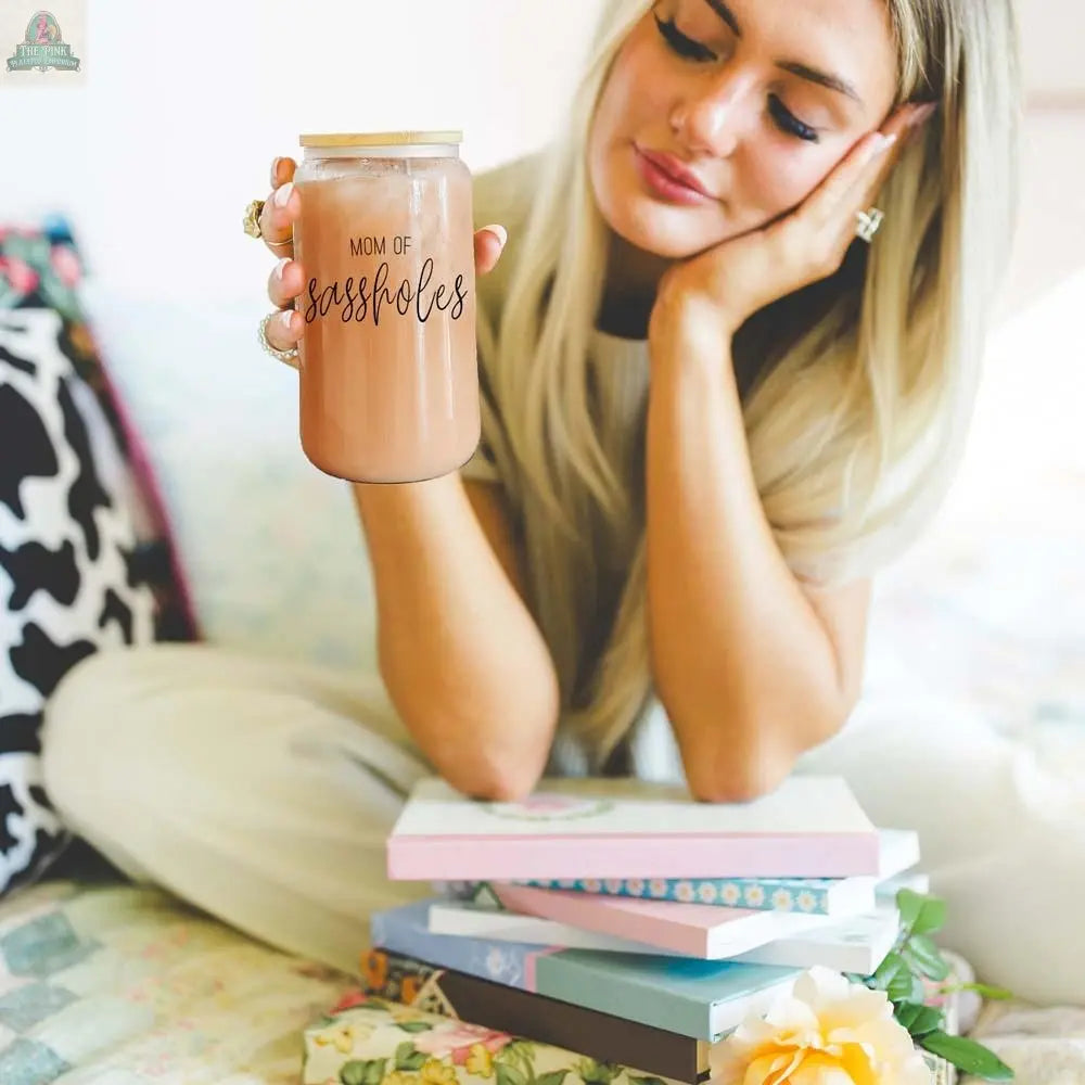 A woman with long blonde hair sits on a bed, holding a Mom of Sassholes 17oz jar. In front of her are glass drinking cups with bamboo lids and stainless steel straws arranged among colorful books and flowers.