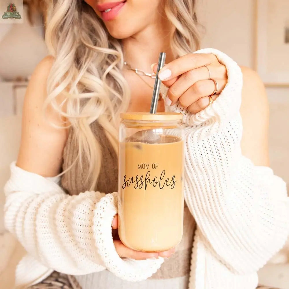 A smiling woman with long, wavy blonde hair holds the "Mom of Sassholes" 17oz glass cup with a stainless steel straw, wearing a white sweater and beige top.