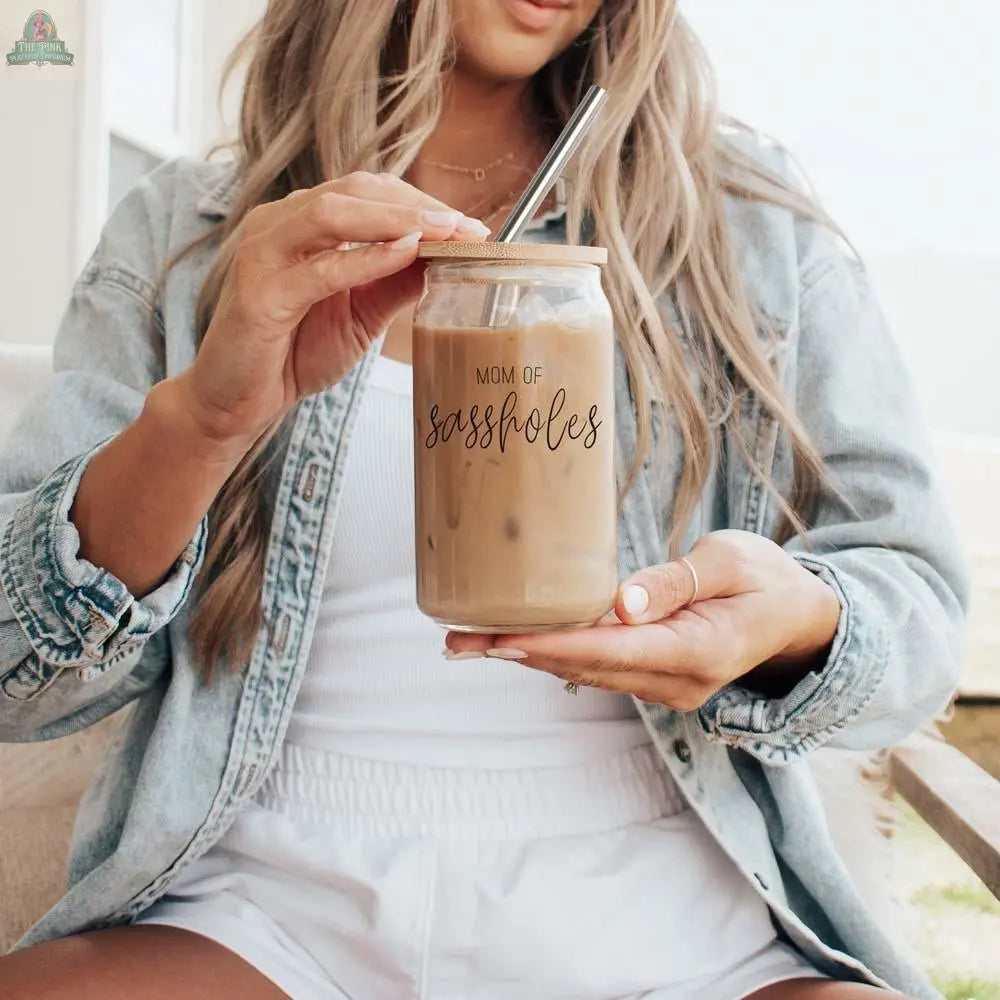 A woman in a light denim jacket and white outfit sits holding an iced coffee in a “Mom of Sassholes 17oz” glass drinking cup with a stainless steel straw.