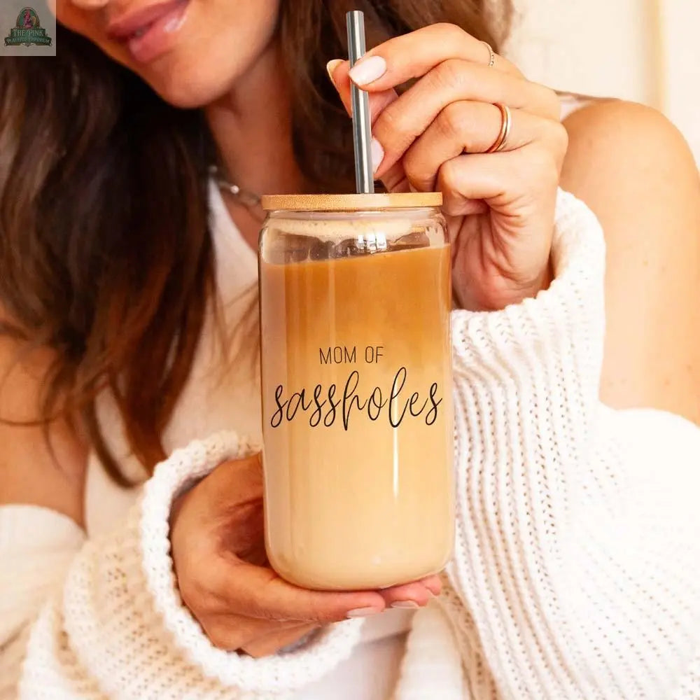 A woman in a white sweater sips iced coffee through a stainless steel straw from the “Mom of Sassholes 17oz” glass, featuring playful black lettering.