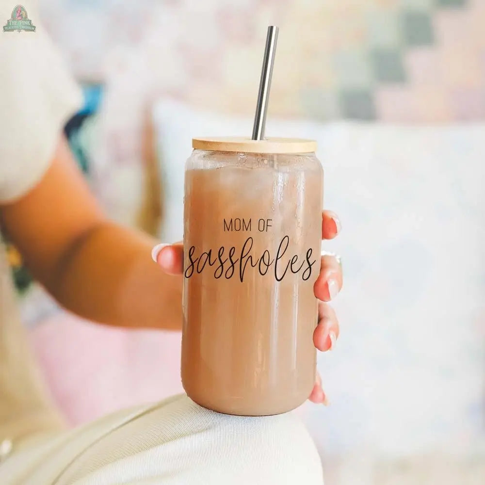 A person holds the Mom of Sassholes 17oz mason jar with iced coffee and a stainless steel straw. “MOM OF sassholes” is written in playful script, while a softly blurred pastel background sets a cheerful mood.