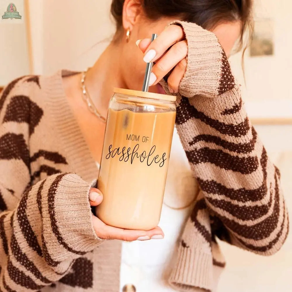 A woman in a brown patterned sweater enjoys iced coffee from the 17oz Mom of Sassholes glass cup, which features a bamboo lid and stainless steel straw, while covering part of her face with one hand.
