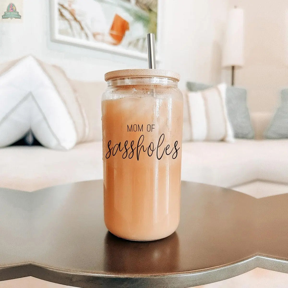 A 17oz Mom of Sassholes glass jar with a stainless steel straw and bamboo lid sits on a coffee table, standing out against a beige couch with white and patterned pillows in the background.