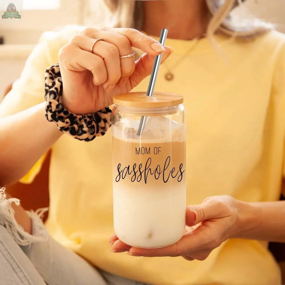 A woman in a yellow shirt holds a "Mom of Sassholes 17oz" glass cup with bamboo lid and stainless steel straw. She wears a leopard print scrunchie and has one knee bent up.