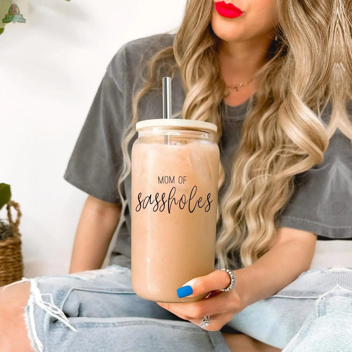 A woman with long blonde hair and red lipstick holds a “Mom of Sassholes” 17oz iced coffee cup with a bamboo lid and stainless steel straw. She wears blue/white nails, a grey t-shirt, and ripped jeans.