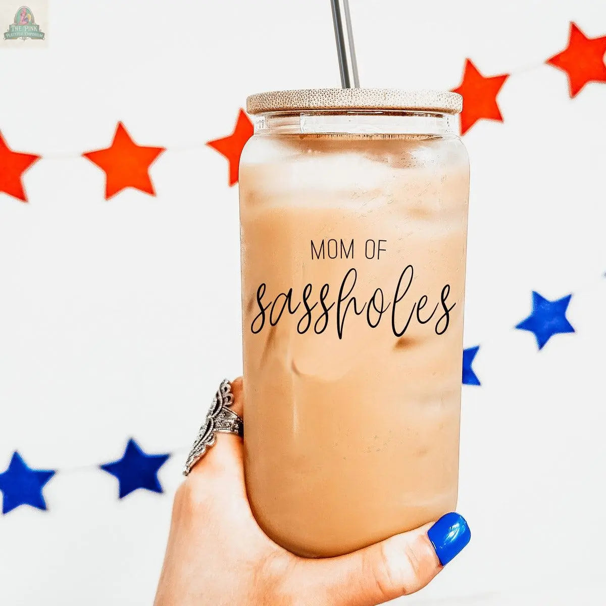 A hand with blue nail polish holds the 17oz "Mom of Sassholes" glass cup with a bamboo lid, filled with iced coffee. A red and blue star garland decorates the white wall in the background.