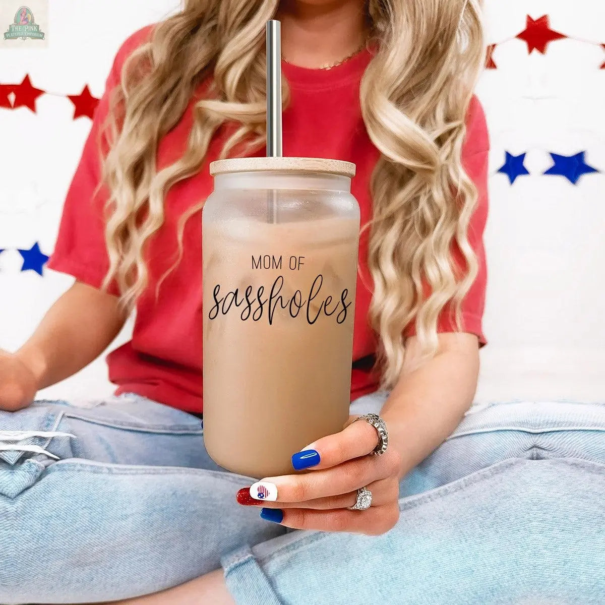A woman with long blonde hair in a red shirt and ripped jeans holds the “Mom of Sassholes 17oz” glass cup with bamboo lid. Her nails are red, white, and blue; star decorations hang in the background.