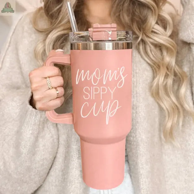 A woman with wavy blonde hair holds the Mom's Sippy 40oz tumbler, featuring a handle and straw. The pink cup with "Mom's Sippy Cup" in white script adds a playful touch to her beige sweater and gold rings.