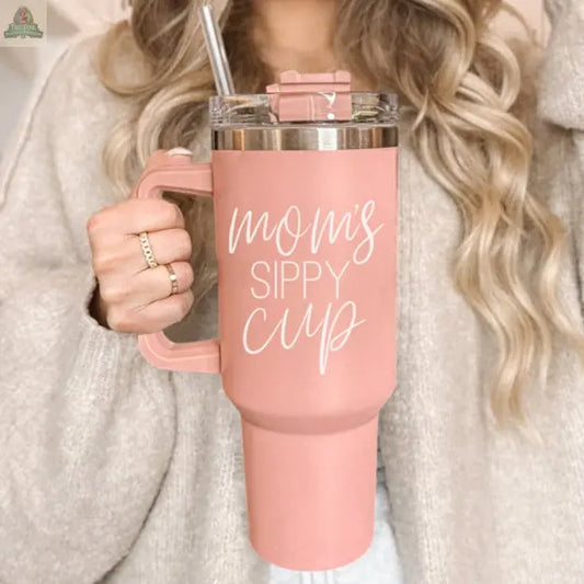 A woman with wavy blonde hair holds the Mom's Sippy 40oz tumbler, featuring a handle and straw. The pink cup with "Mom's Sippy Cup" in white script adds a playful touch to her beige sweater and gold rings.