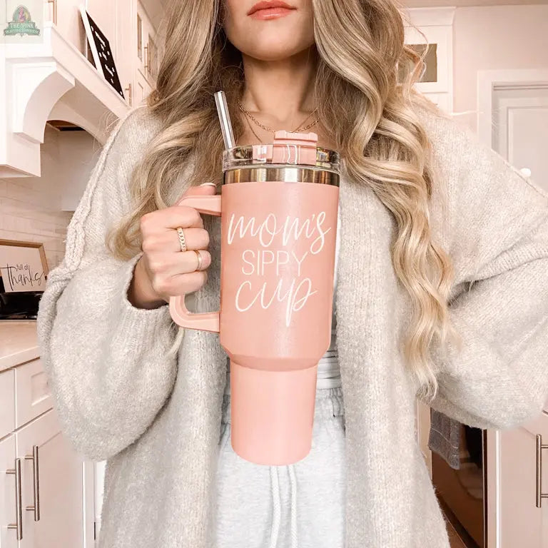 A woman with wavy blonde hair wears a cozy beige cardigan and holds a Mom's Sippy 40oz tumbler in a bright, modern kitchen.