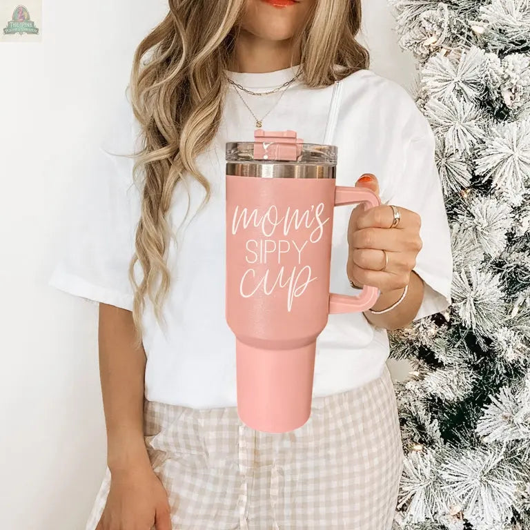 A woman with long wavy hair holds a pink Mom's Sippy 40oz tumbler near a frosted Christmas tree, wearing a white shirt and plaid pants.