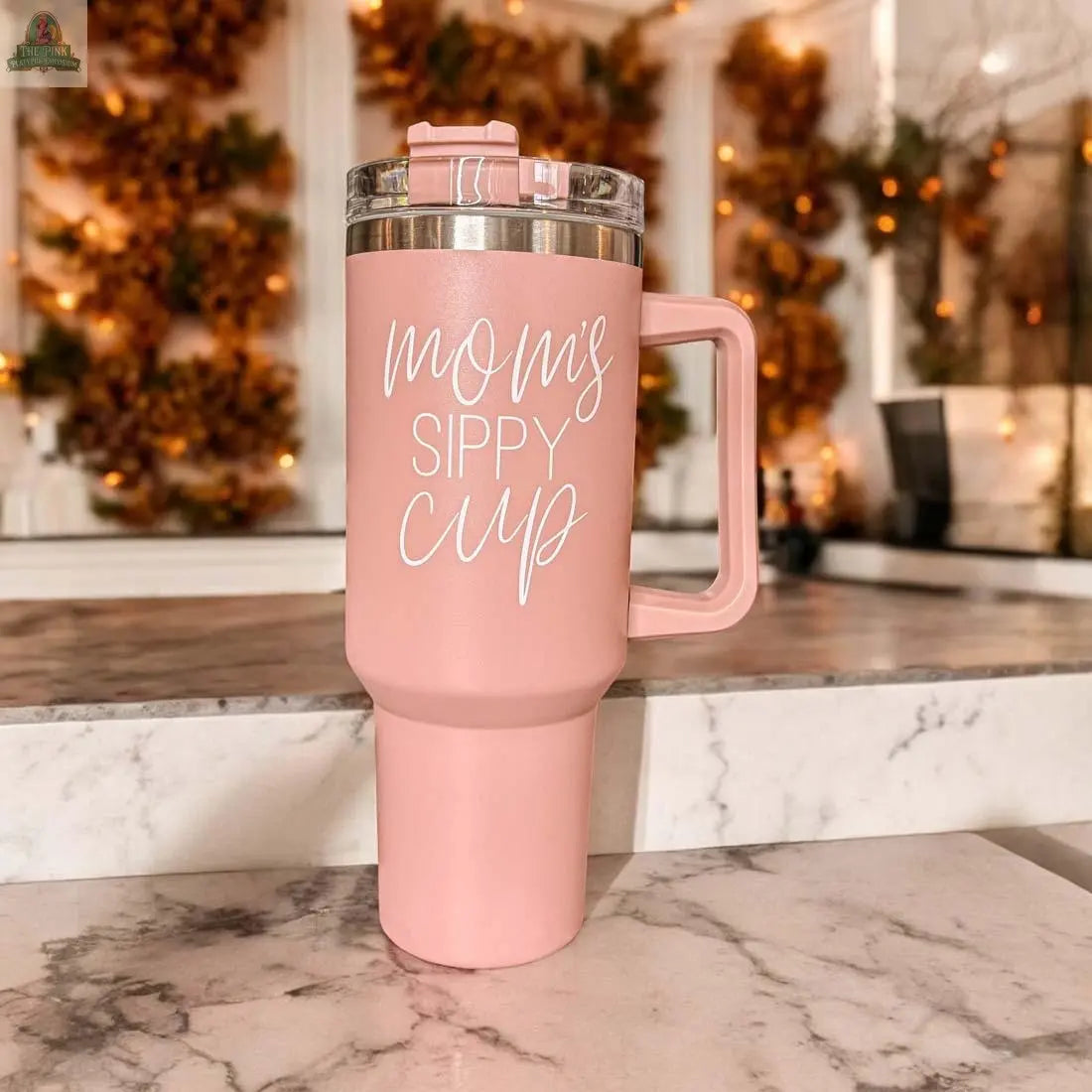 The Mom’s Sippy 40oz, a pink tumbler with a handle and clear lid, features “Mom’s Sippy Cup” in white script. Set on marble with soft lighting and autumn leaves, it’s perfect for cozy days.