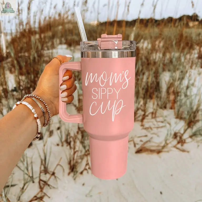 A hand holds the Mom's Sippy 40oz, a large pink tumbler with a handle, straw, and lid. "Mom's Sippy Cup" is written in white script. A sandy beach and tall grasses appear in the background.