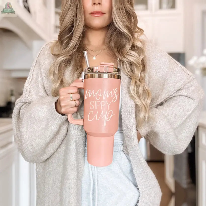 A woman with long, wavy blonde hair stands in a kitchen holding the Mom's Sippy 40oz tumbler with a straw. She wears a beige cardigan and light blue pants.