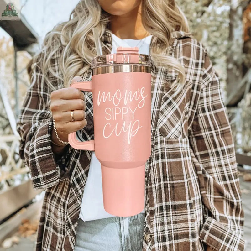 A woman in a plaid shirt holds the Mom's Sippy 40oz tumbler in pink with white script. Trees and outdoor scenery appear in the background.