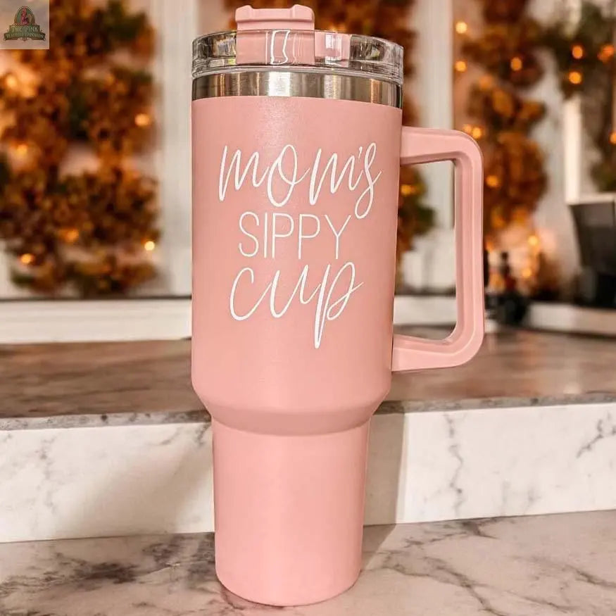A large pink Mom's Sippy 40oz tumbler with handle and lid, featuring “Mom’s Sippy Cup” in white script, sits on a marble counter, blurred autumn decorations in the background.