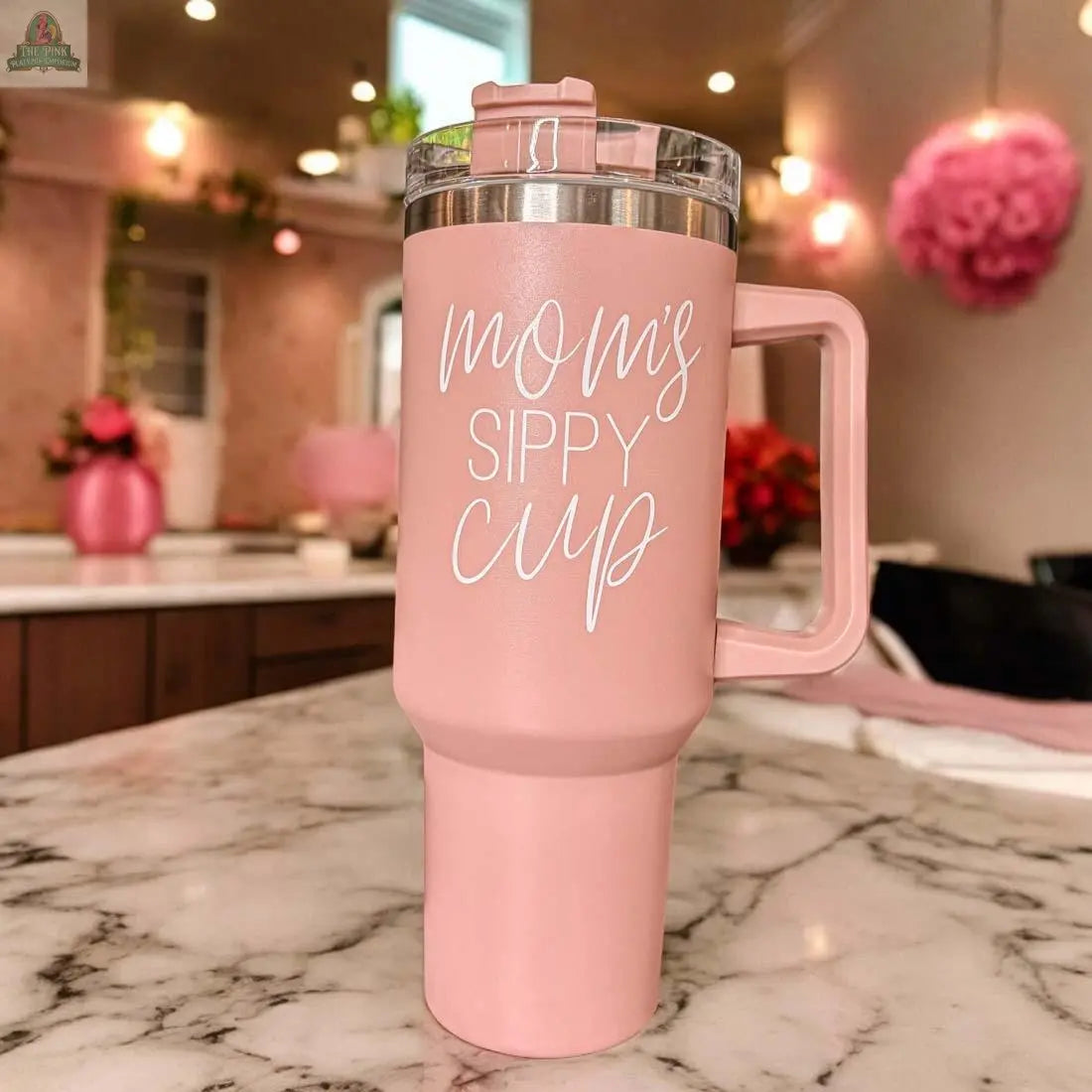 The Mom’s Sippy 40oz tumbler is a pink mug with a handle and clear lid, featuring “mom’s SIPPY CUP” in white script. Set on a marble counter in a pink-accented kitchen, it’s a perfect addition to any mom tumbler collection.