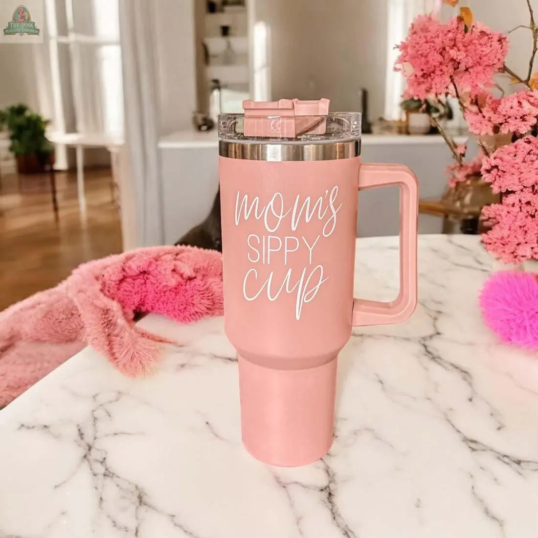 The Mom's Sippy 40oz tumbler in pink with a handle rests on a marble table, displaying “mom’s sippy cup” in white script. Pink flowers and soft decorations enhance the scene in the background.