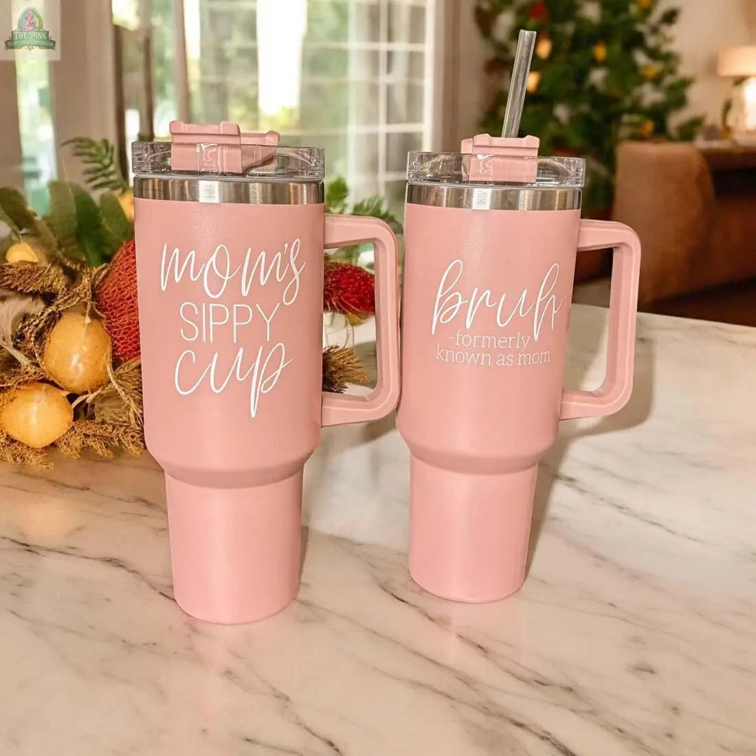 Two pink Mom's Sippy 40oz tumblers with handles rest on a marble countertop. One tumbler reads "Mom's Sippy Cup," while the other says "bruh – formerly known as mom." Blurred holiday decor appears in the background.