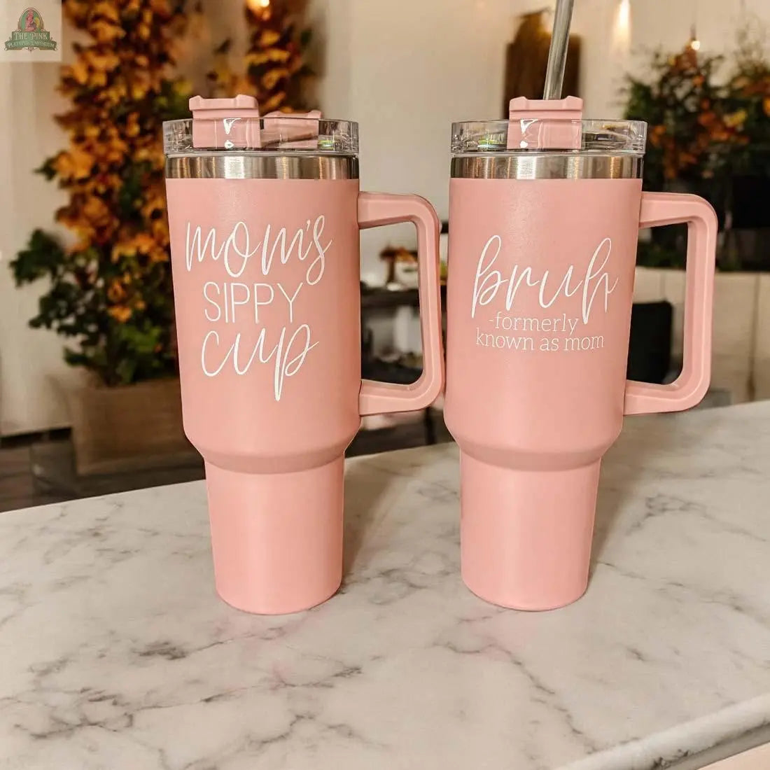 Two pink Mom's Sippy 40oz tumblers with handles rest on a marble counter. One says "Mom's Sippy Cup," the other reads "bruh - formerly known as mom." Both have pink lids, with fall decor in the background.