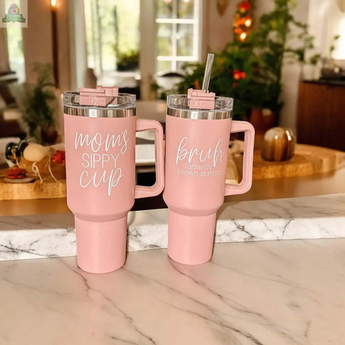 Two Mom's Sippy 40oz pink tumblers rest on a marble countertop; one features “mom’s SIPPY CUP” while the other says “bruh (formerly known as mom),” with a cozy, plant-filled kitchen in the background.
