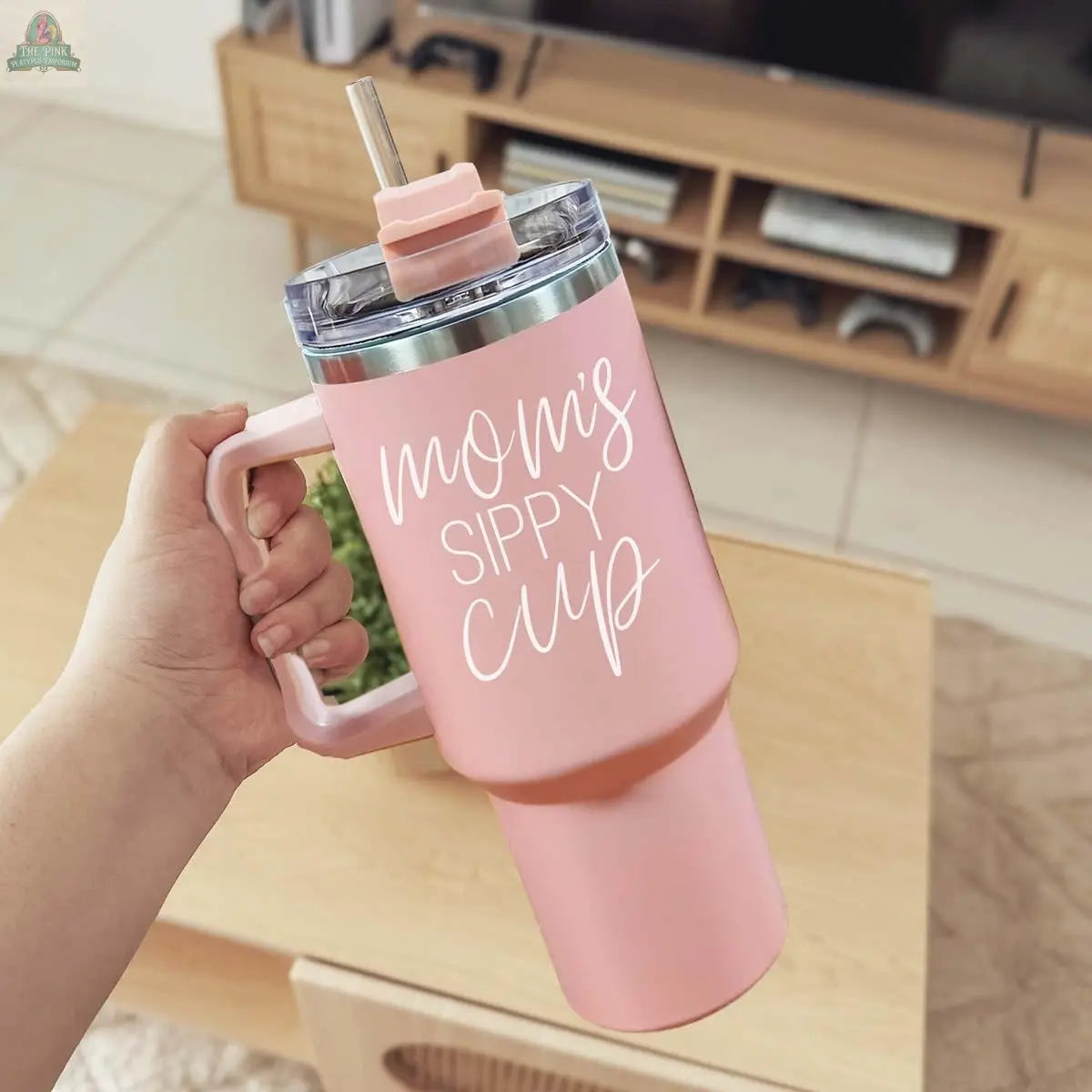 A hand holds the Mom's Sippy 40oz tumbler with a straw and lid, featuring "Mom's Sippy Cup" in white cursive. A wooden TV stand and tiled floor appear in the background.