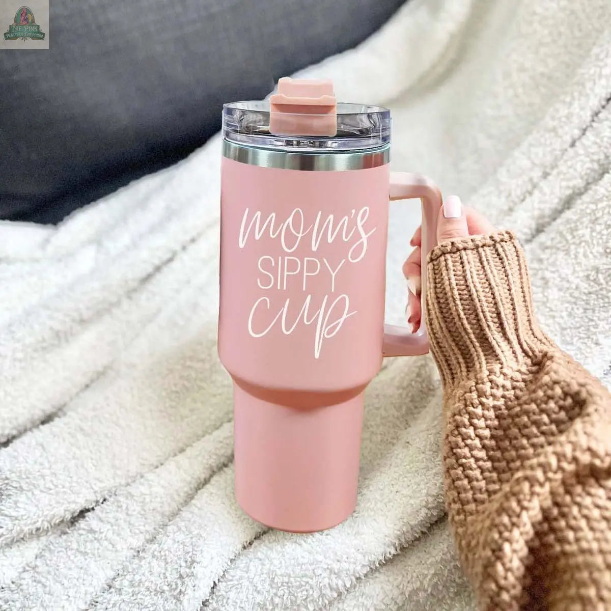 A hand in a brown knit sweater holds the Mom’s Sippy 40oz pink tumbler with lid, featuring “Mom’s Sippy Cup” in white script, against a cozy white fuzzy blanket background.