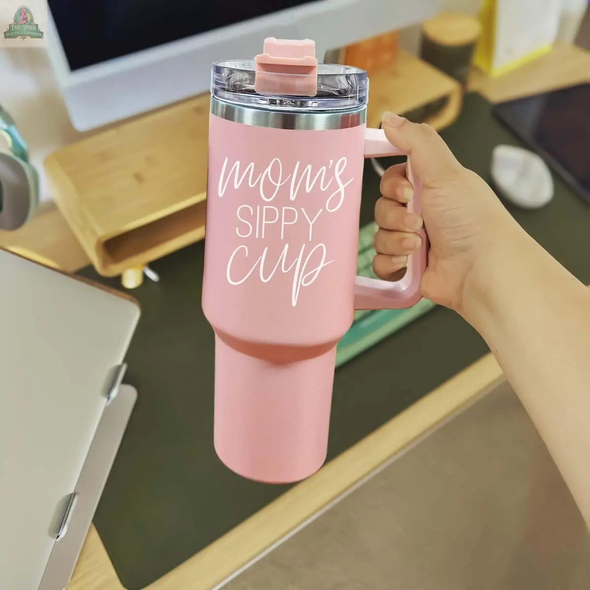 A hand holds the Mom's Sippy 40oz tumbler with "Mom's Sippy Cup" in white script, set against a desk with a computer, keyboard, and office essentials in the background.