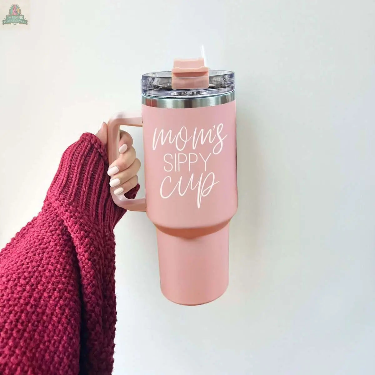 A hand in a red knit sweater holds the Mom's Sippy 40oz tumbler—large, pink, with a straw and white “Mom's Sippy Cup” lettering.