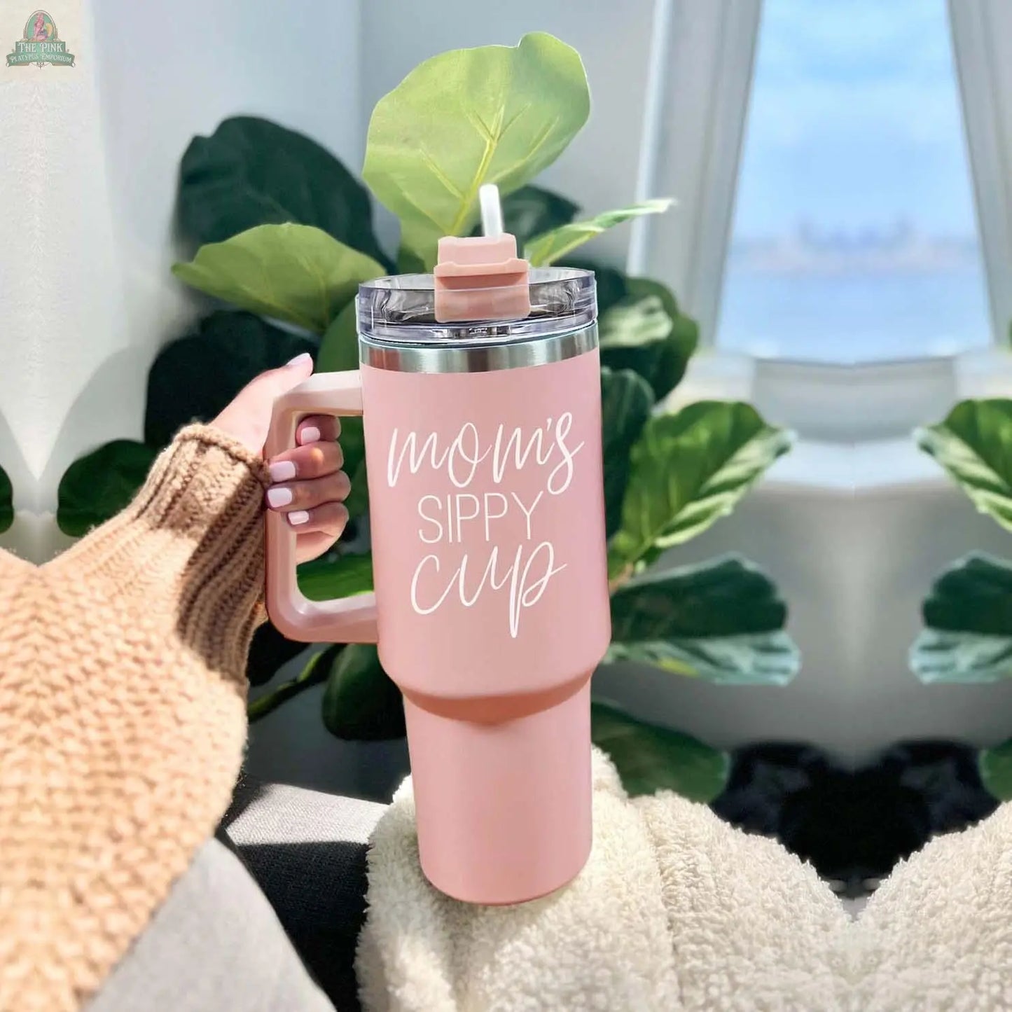 A person in a cozy sweater holds the Mom's Sippy 40oz Tumbler with lid and straw, sitting by green plants near a window with a city view.