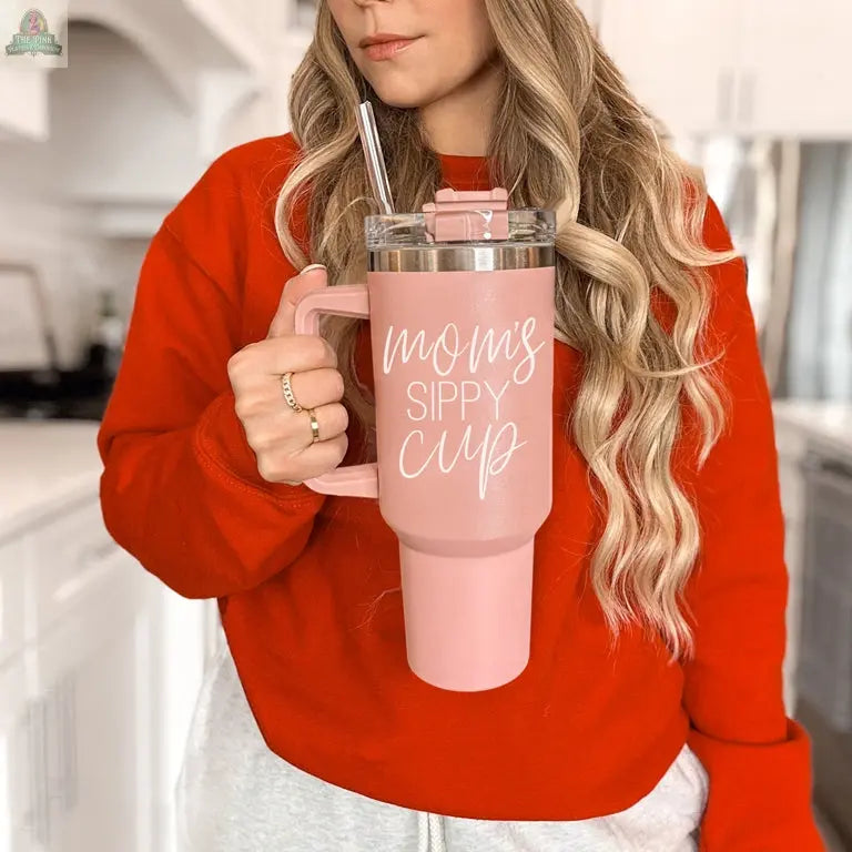 A woman in a red sweatshirt holds the bright pink Mom's Sippy 40oz tumbler with lid and straw, featuring “Mom's Sippy Cup” in white script, while standing in a cheerful, sunlit kitchen.