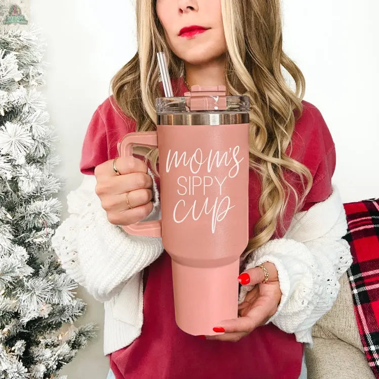 A woman in a pink top and white cardigan holds a Mom's Sippy 40oz tumbler with lid and straw. A decorated white Christmas tree stands beside her.