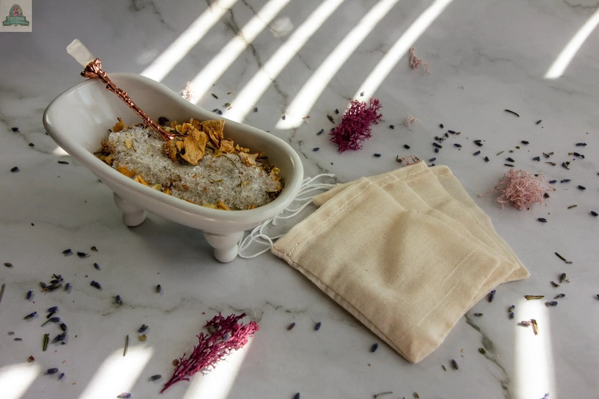 Moon Magic features a small white bathtub dish with jasmine and vanilla bath salts, dried flowers, and a scoop. Three beige fabric pouches of herbal bath tea sit on marble amid scattered petals, illuminated by soft sunlight.