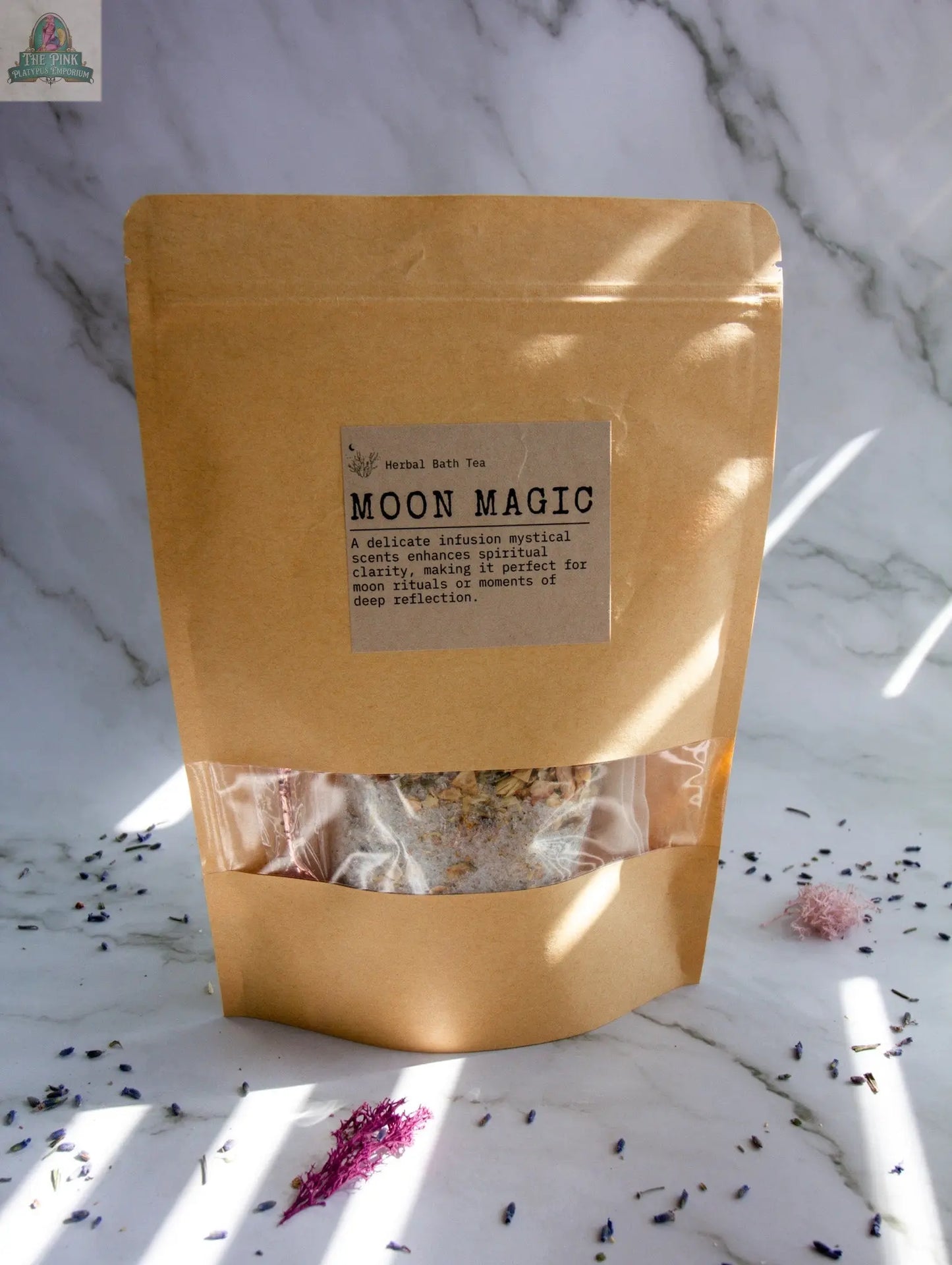 A brown kraft paper pouch labeled “Moon Magic” sits on a marble surface with sunlight and flower petals nearby. The pouch’s clear window displays dried herbs infused with jasmine and vanilla essential oils.