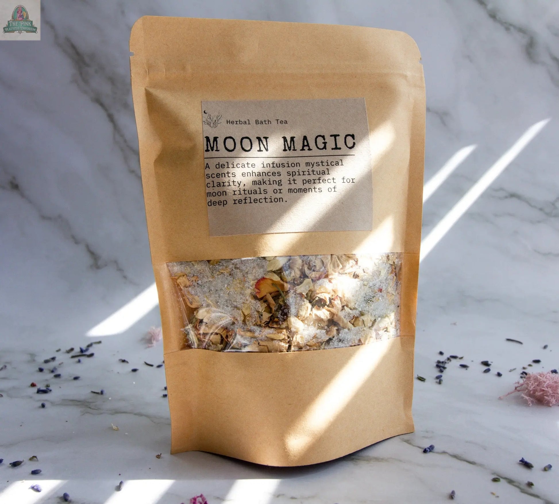 A kraft paper bag labeled “Moon Magic” stands upright on a marble background. Dried herbs and flowers for moon rituals are visible through a clear window. The label notes the bath tea enhances spiritual clarity.
