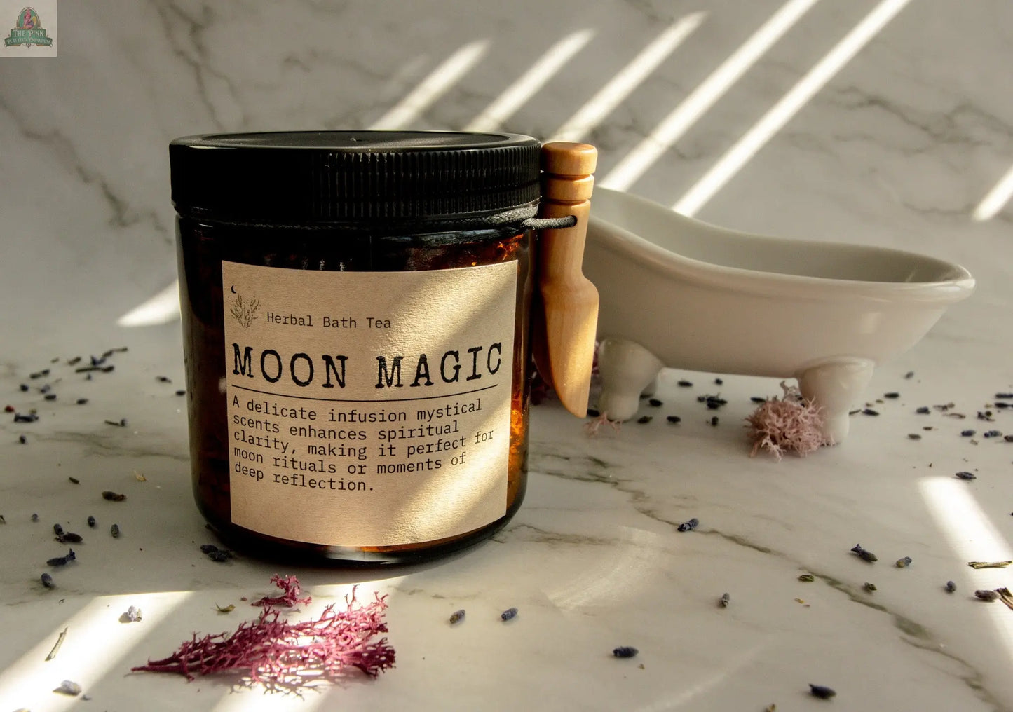 A jar labeled Moon Magic with a wooden scoop sits by a white tub; dried flower petals—ideal for moon rituals—are scattered on marble, illuminated by sunlight and shadow patterns.