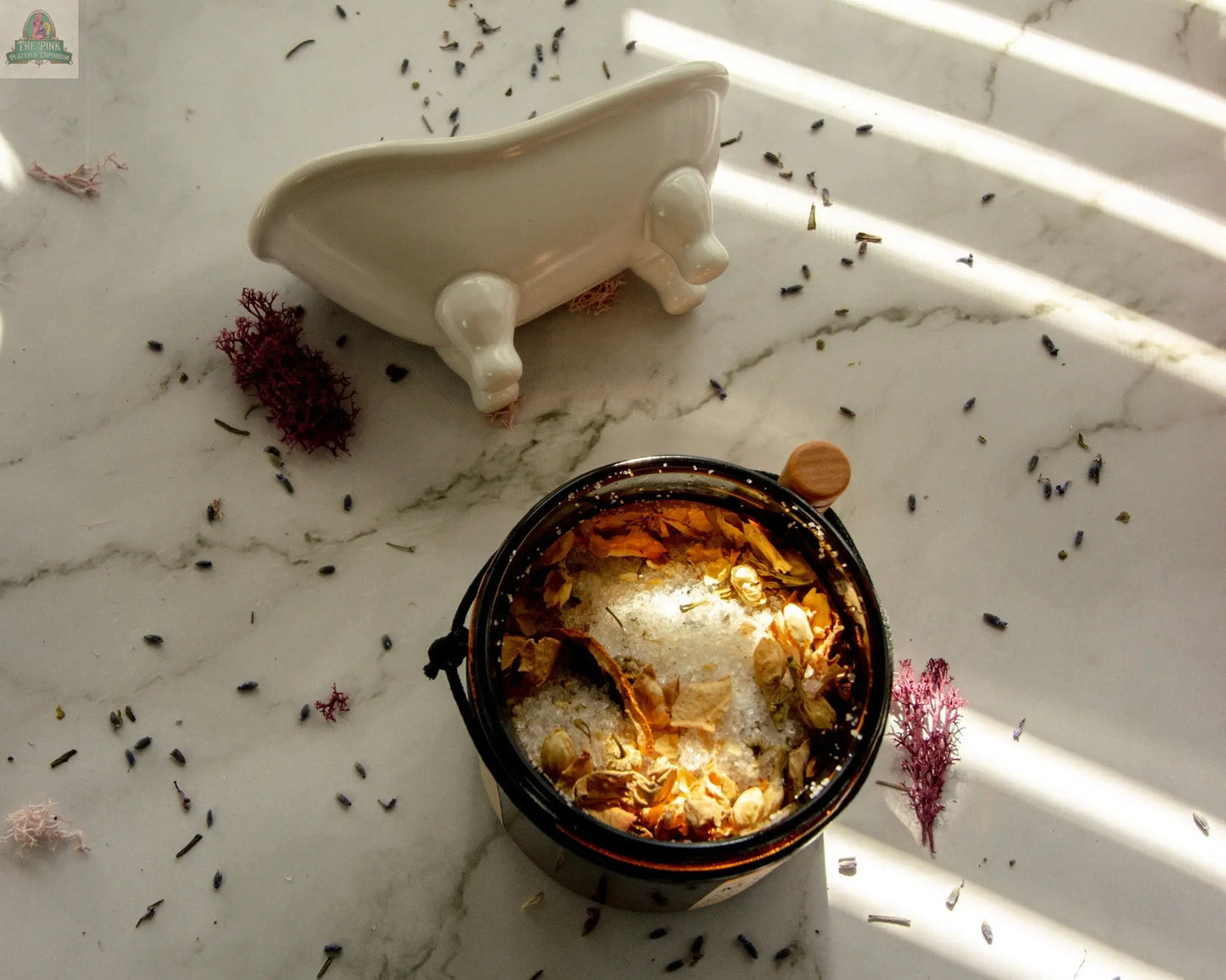 A jar of Moon Magic bath salts, infused with jasmine and vanilla essential oils, rests on a marble surface scattered with petals, while a small white bathtub dish sits in the background as sunlight casts gentle shadows.