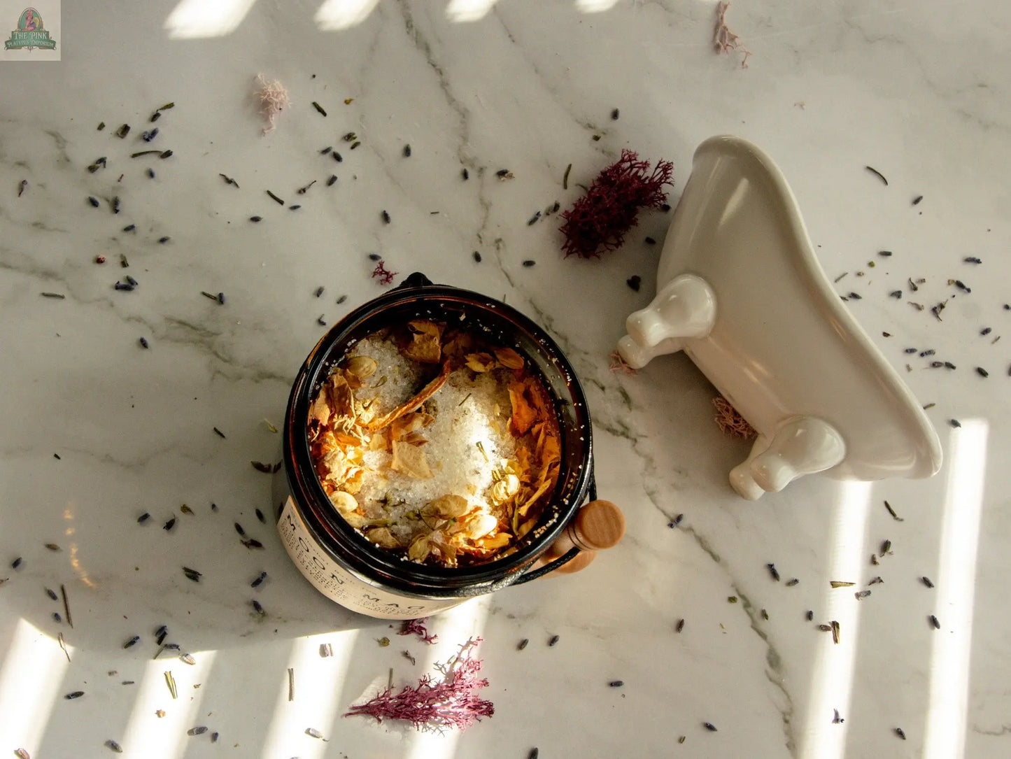 A jar of Moon Magic herbal bath tea, filled with lavender buds and purple dried flowers infused with jasmine and vanilla essential oils, sits on marble beside a white clawfoot tub dish, with flowers scattered in sunlight.