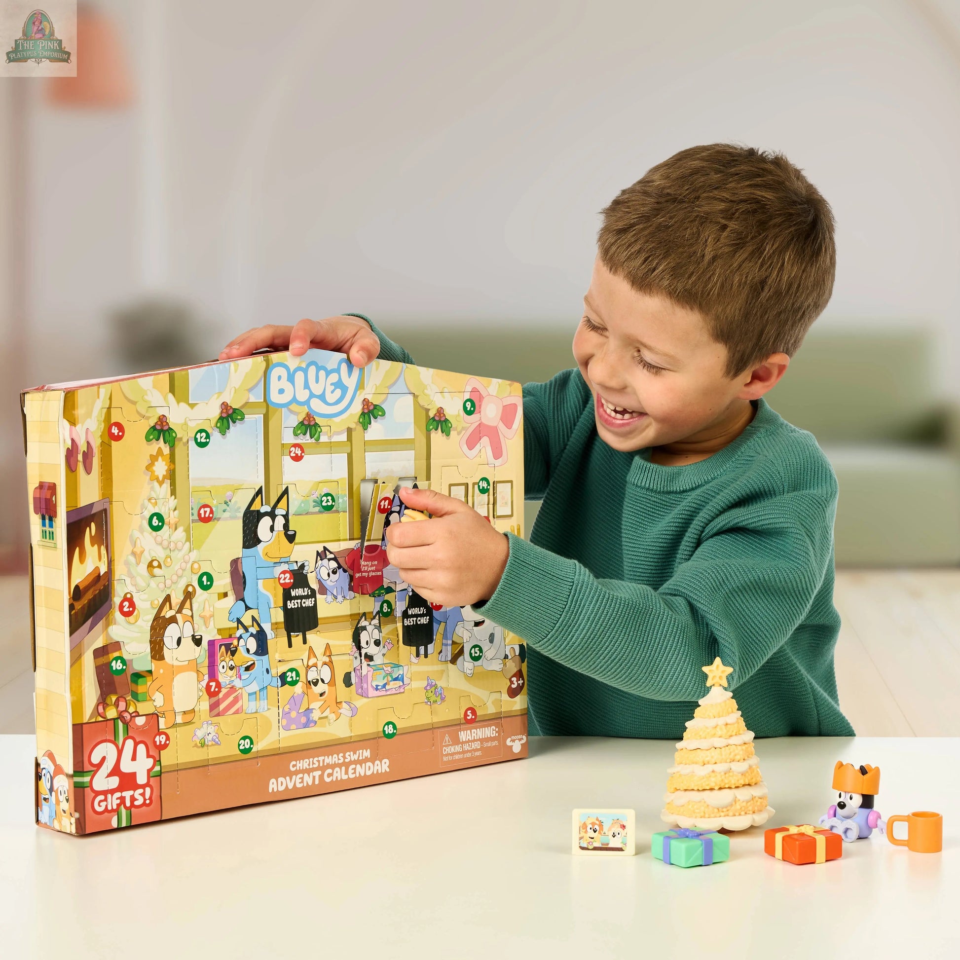 A young boy smiles as he opens the Bluey Advent Calendar, which features cartoon characters and festive designs. A decorated Christmas tree and small presents sit on the table, making each day a fun Bluey surprise.