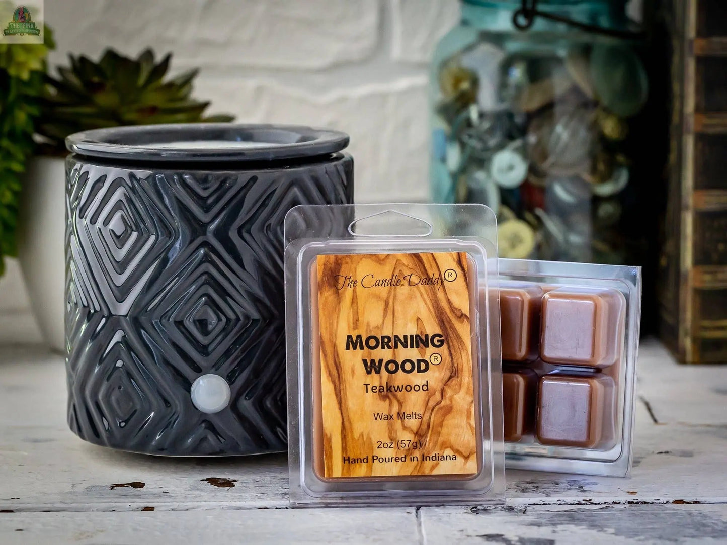 A gray patterned wax warmer sits on a white surface next to Candle Daddy’s Morning Wood - Teak Wood Scented Wax Melt, with one labeled pack and another open showing brown cubes. Decorative items are blurred in the background.