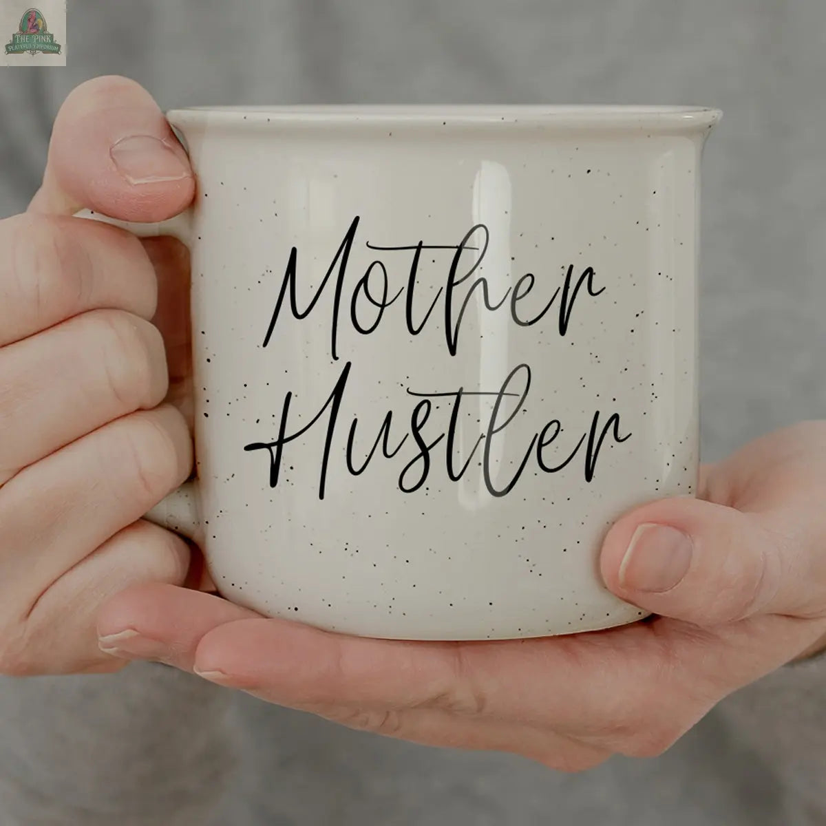 A person in a light grey top holds the Mother Hustler 14.5oz mug, a white speckled coffee mug with black cursive text—perfect as a motivational gift for any mom boss.