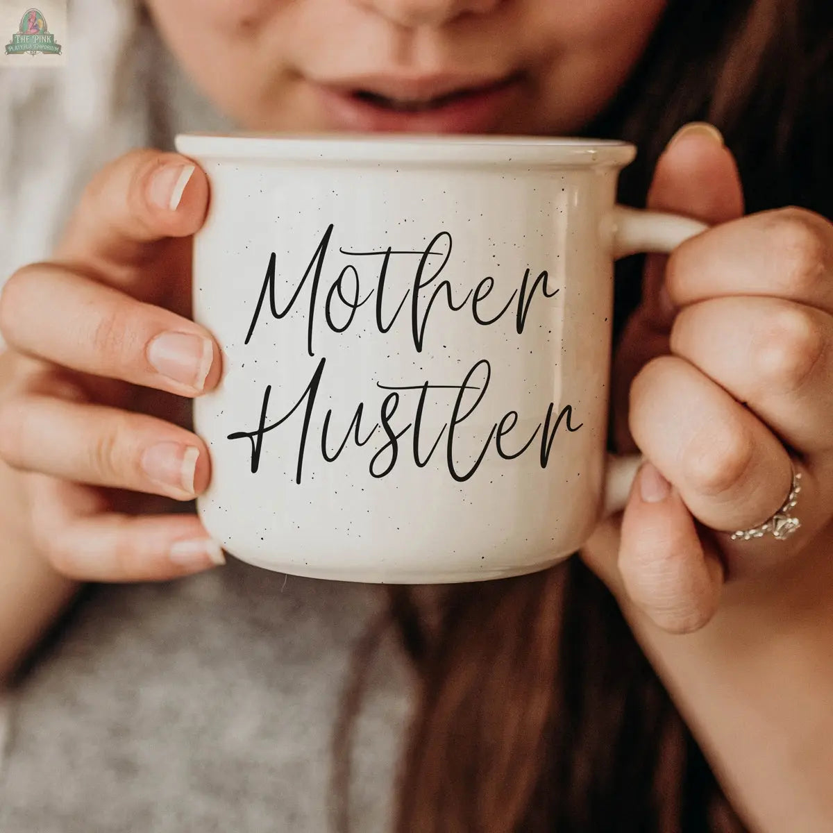 A woman with long brown hair holds the 14.5oz Mother Hustler mug close to her face. Wearing a gray top and a ring, she captures a true mom boss mug moment, with "Mother Hustler" in bold black script on her coffee mug.