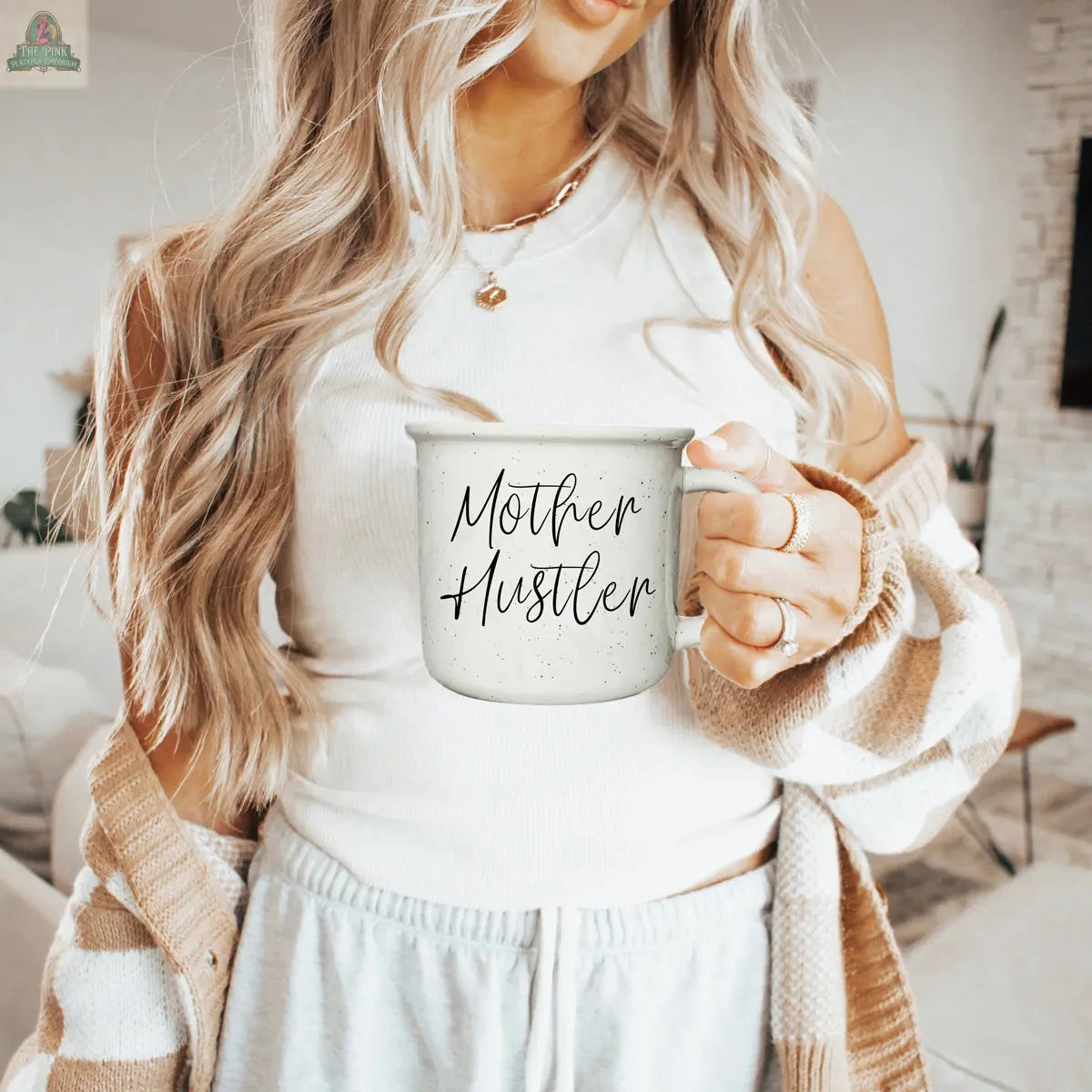 A woman with long blonde hair in a white tank top and cardigan holds the Mother Hustler 14.5oz mug while standing in a stylish, softly-lit living room.