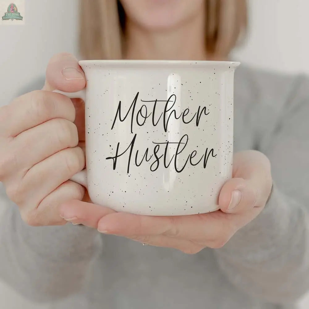A person holds the Mother Hustler 14.5oz mug, a white speckled cup with black cursive lettering. Wearing a light gray top, only their lower face and hands are visible, highlighting this motivational mug for any mom boss.