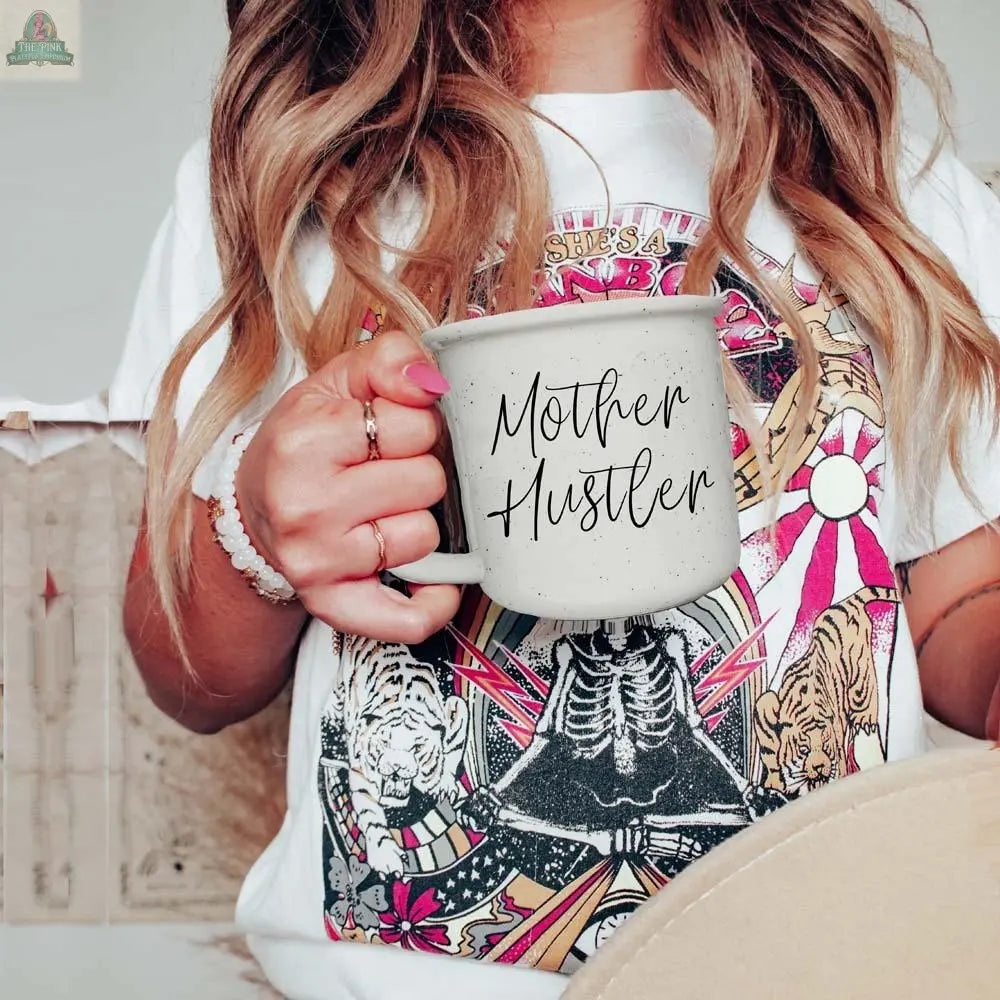 A woman with long wavy hair holds a Mother Hustler 14.5oz coffee mug. She wears a colorful graphic tee featuring tigers and skeletons, accessorized with painted nails, a bracelet, and a ring.