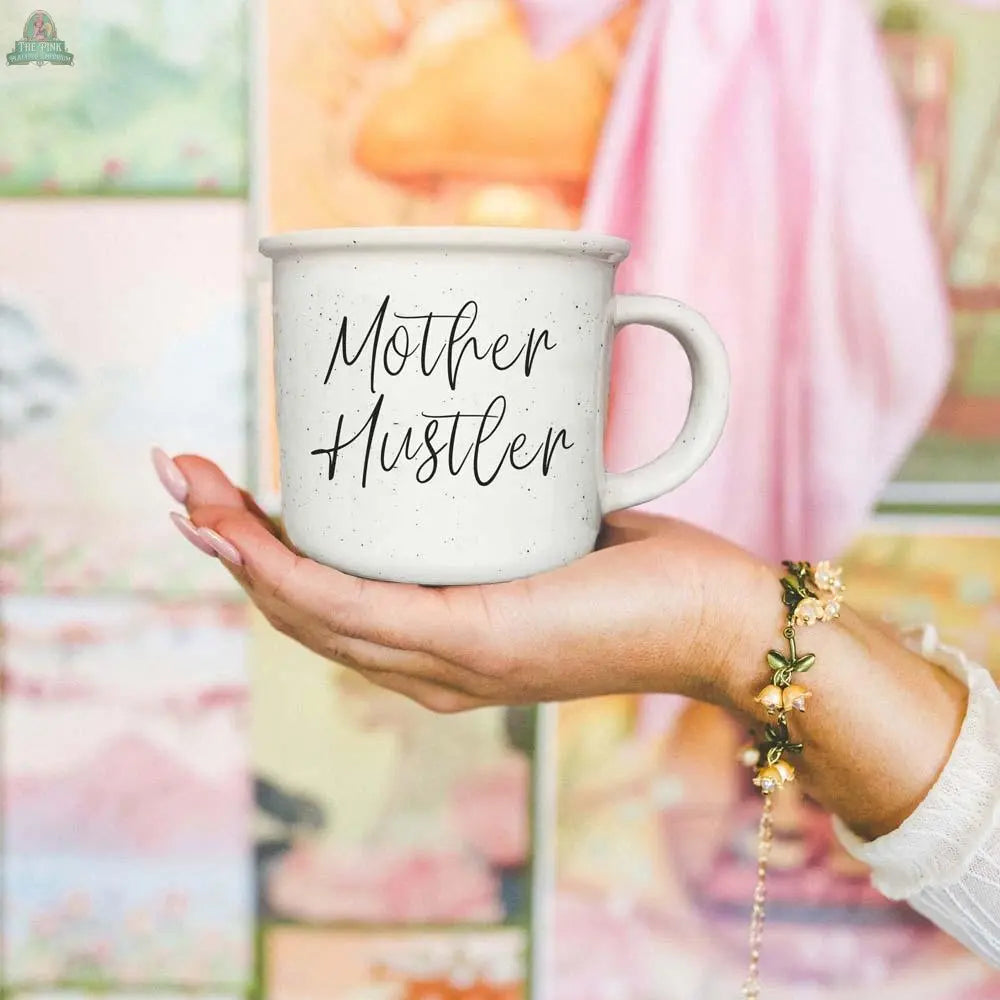 A hand with a floral bracelet holds the Mother Hustler 14.5oz mug, featuring cursive lettering, with vibrant wall art softly blurred in the background.