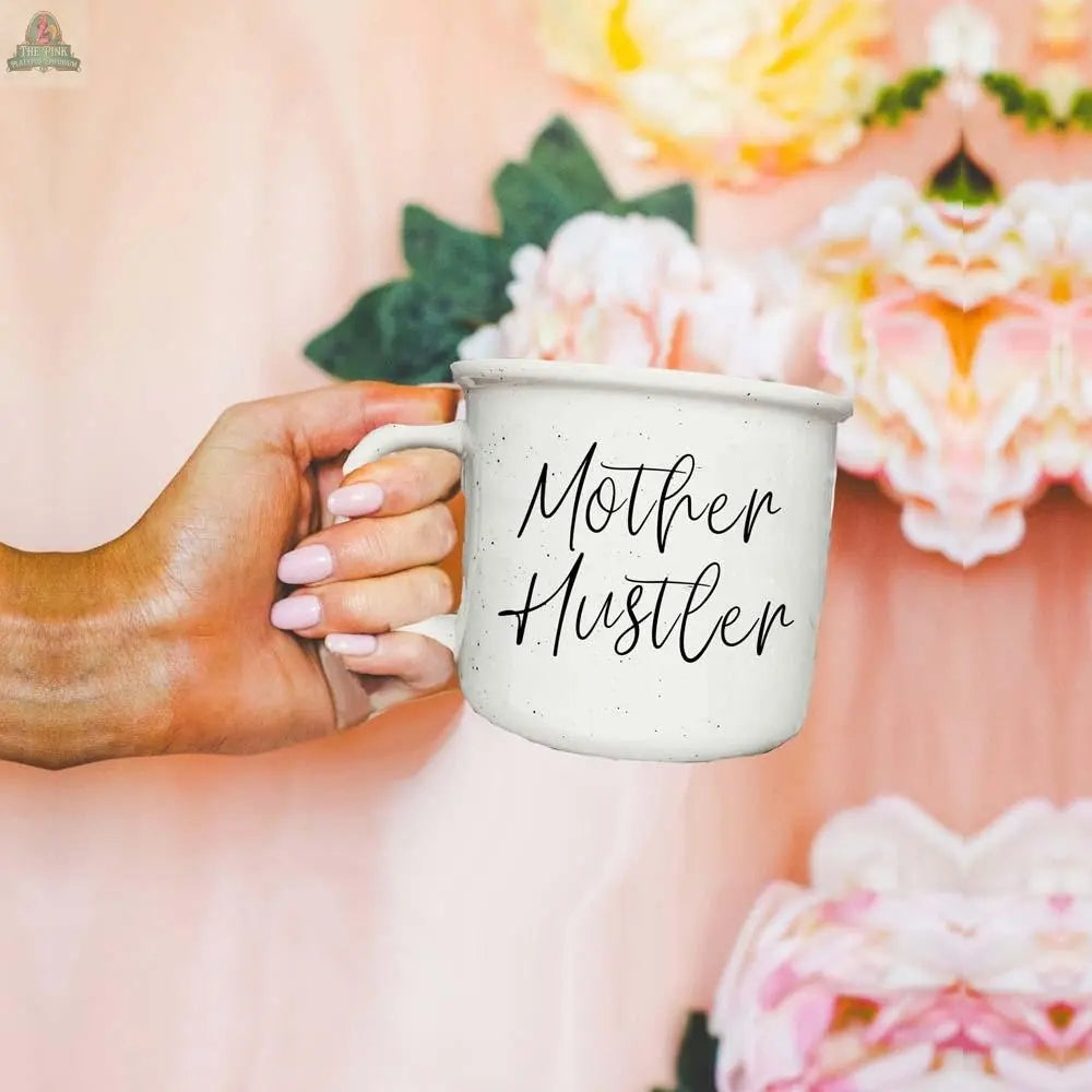 A hand with light pink nails holds the Mother Hustler 14.5oz mug, with soft pink and yellow flowers blurred in the background. This motivational mug inspires confidence and hustle with every sip.