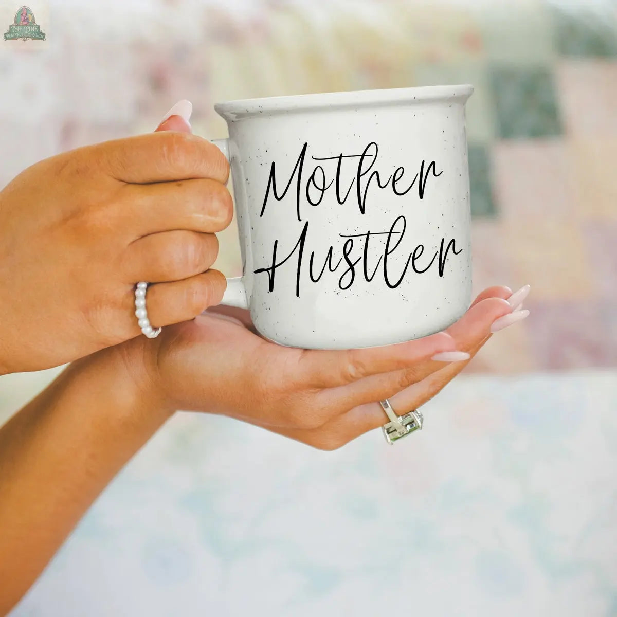 A person with manicured nails and rings holds the Mother Hustler 14.5oz white speckled mug with cursive text, against a softly blurred pastel background.