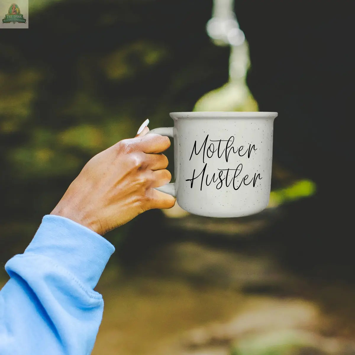 A person in a light blue sleeve holds the Mother Hustler 14.5oz mug with bold lettering. The blurred outdoor background makes this motivational coffee mug the perfect accessory for any go-getter mom.